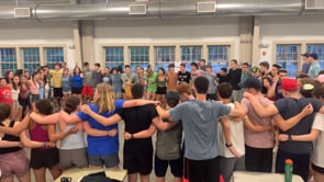 A large group of people standing in a circle indoors with their arms around each other’s shoulders, appearing to participate in a group activity or gathering.