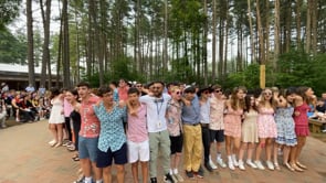 A large group of teens dressed in summer clothes stand arm in arm, smiling outdoors on a brick path, with tall trees and an audience seated in the background.