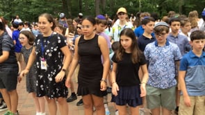 A group of children and teens stand outdoors in a crowded area, some dressed in casual summer clothing, surrounded by trees and greenery. They appear attentive and ready for an activity.
